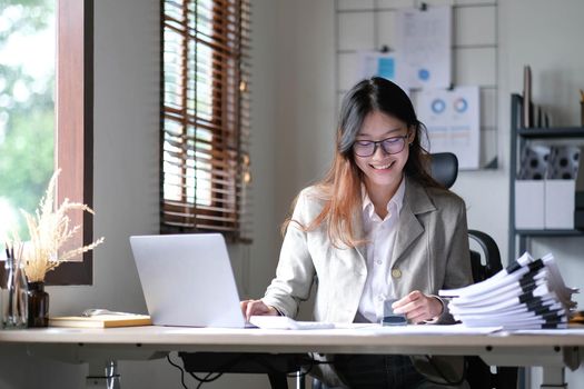 Beautiful Asian businesswoman working on a desk with a laptop with a smiling face while looking at the document received in the office..