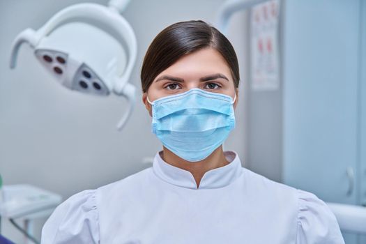Headshot portrait of young female doctor nurse dentist looking at camera in medical face mask in white, dental cabinet with equipment. Dentistry medicine, specialist career, dental health care concept