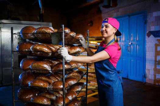 Professional baker - a young, pretty woman in a jeans apron holds fresh bread against the background of a bakery or bakery. Bakery products. Bread production