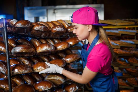 A young girl works in a bakery. She puts bread on a shelf. Woman baker at workplace in a bakery. A professional baker holds bread in his hands. Bread production concept