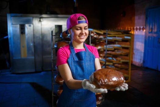 A baker girl takes hot bread in a bakery against the background of shelving with bread. Industrial production of bread. The stage of baking in a bakery