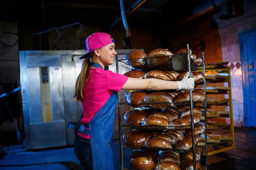 Professional baker - a young, pretty woman in a jeans apron holds fresh bread against the background of a bakery or bakery. Bakery products. Bread production