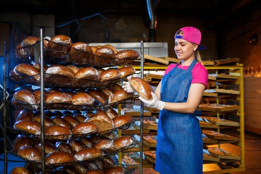 Professional baker - a young, pretty woman in a jeans apron holds fresh bread against the background of a bakery or bakery. Bakery products. Bread production