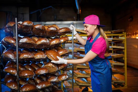Professional baker - a young, pretty woman in a jeans apron holds fresh bread against the background of a bakery or bakery. Bakery products. Bread production
