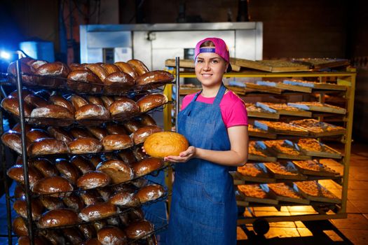 Professional baker - a young, pretty woman in a jeans apron holds fresh bread against the background of a bakery or bakery. Bakery products. Bread production