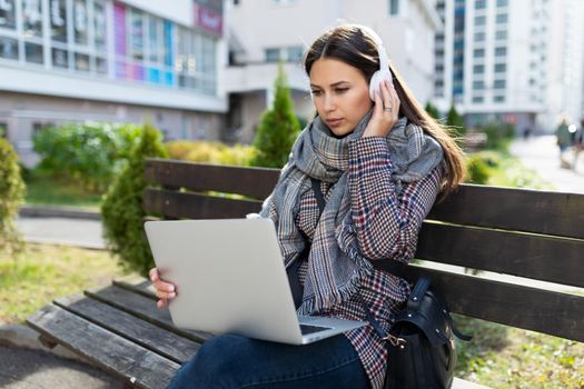 a young woman works online on a laptop and listens to music on headphones while sitting on a bench next to a shopping center.