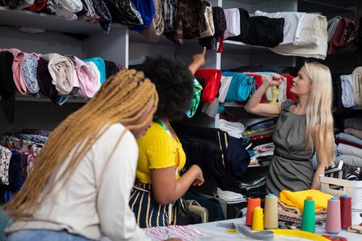 A dark-skinned shopkeeper shows a blonde woman what materials are most appropriate for a summer dress. Two African women and a European woman work in a tailor's shop.