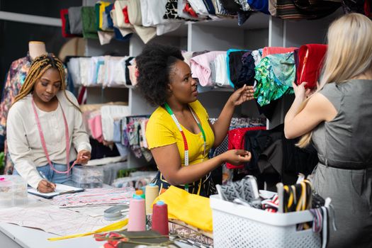 A dark-skinned shopkeeper shows a blonde woman what materials are most appropriate for a summer dress. Two African women and a European woman work in a tailor's shop.