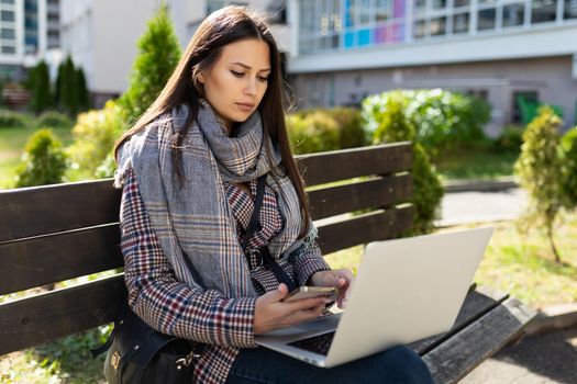 a young woman in autumn clothes is working intently on a laptop while sitting on a bench outside.