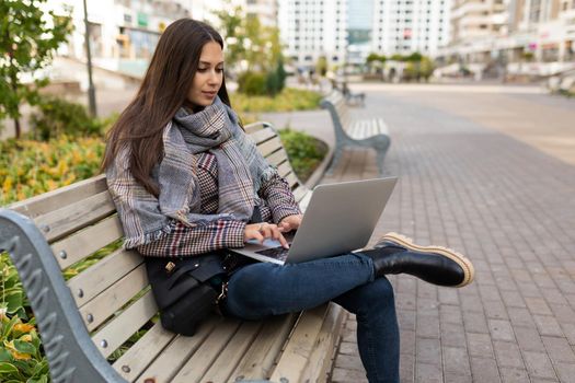 young successful female freelancer working on a laptop against the backdrop of a city park sitting in warm autumn clothes.