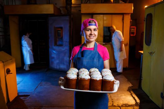 Girl baker holds a tray with hot pastries in the bakery. She is wearing a denim frock and a cap. Production of bakery products. Rack with fresh crispy pastries.