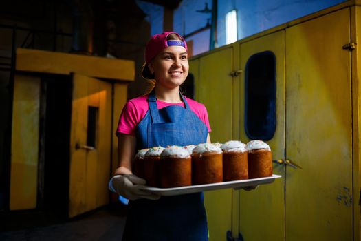 Girl baker holds a tray with hot pastries in the bakery. She is wearing a denim frock and a cap. Production of bakery products. Rack with fresh crispy pastries.