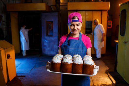 Girl baker holds a tray with hot pastries in the bakery. She is wearing a denim frock and a cap. Production of bakery products. Rack with fresh crispy pastries.