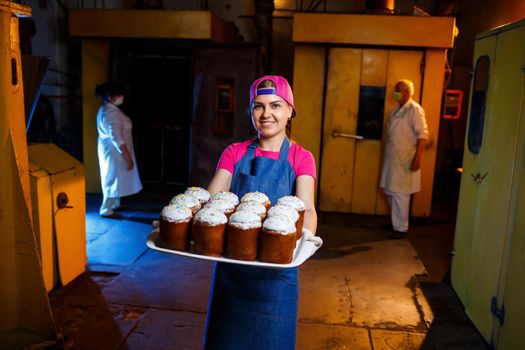 Girl baker holds a tray with hot pastries in the bakery. She is wearing a denim frock and a cap. Production of bakery products. Rack with fresh crispy pastries.