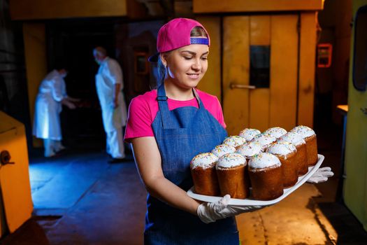 Girl baker holds a tray with hot pastries in the bakery. She is wearing a denim frock and a cap. Production of bakery products. Rack with fresh crispy pastries.