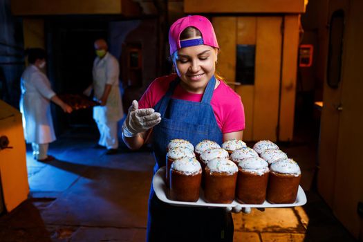 Girl baker holds a tray with hot pastries in the bakery. She is wearing a denim frock and a cap. Production of bakery products. Rack with fresh crispy pastries.