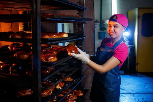 Girl baker holding fresh pies in her hands while standing in the bakery. She is wearing a denim apron and a cap. Production of bakery products. Shelf with hot crispy pastries.