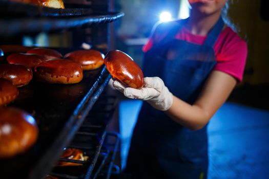 Girl baker holds fresh pies in the hands of the bakery. She is wearing a denim frock and a cap. Production of bakery products. Rack with hot crispy pastries.