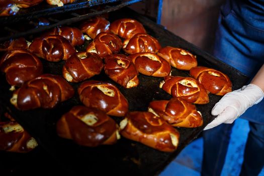 Girl baker holds a tray with hot pastries in the bakery. She is wearing a denim frock and a cap. Production of bakery products. Rack with fresh crispy pastries.