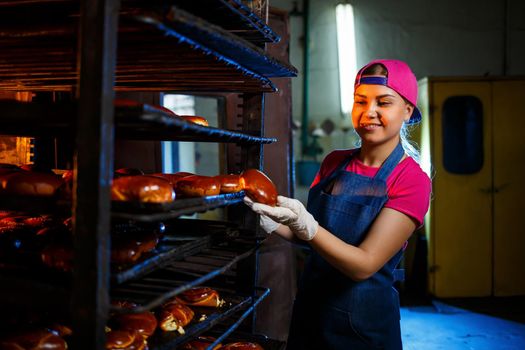 Girl baker holds fresh pies in the hands of the bakery. She is wearing a denim frock and a cap. Production of bakery products. Rack with hot crispy pastries.
