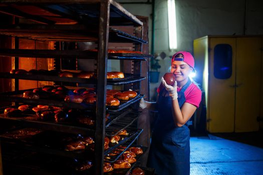 Girl baker holds fresh pies in the hands of the bakery. She is wearing a denim frock and a cap. Production of bakery products. Rack with hot crispy pastries.