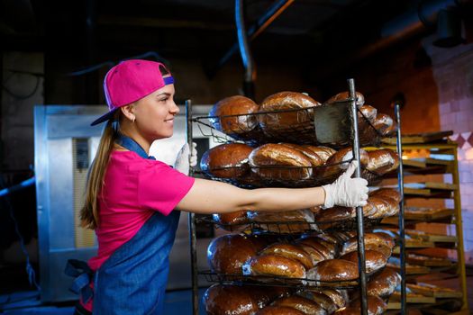 A baker girl takes hot bread in a bakery against the background of shelving with bread. Industrial production of bread. The stage of baking in a bakery