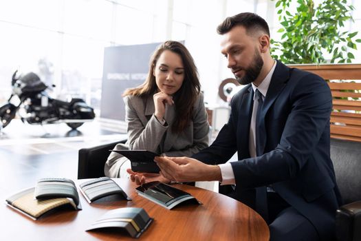 car dealership manager helping young woman choose car trim when buying it at car dealership.