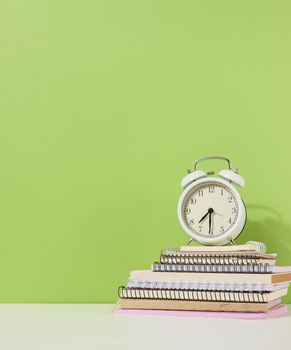 Round alarm clock stands on a stack of notebooks, green background. Back to school