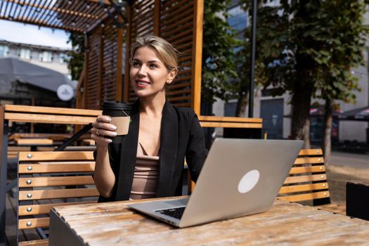 stylish businesswoman drinking coffee and working on terrace with laptop outdoors.