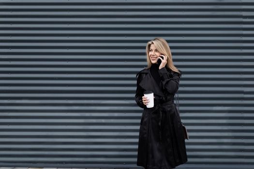 modern businesswoman with a cup of coffee in her hands in business clothes speaks on the phone against the background of a gray textured wall.