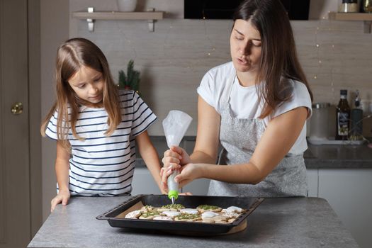 A little girl with her mother decorates Christmas cookies in the kitchen. Real life.