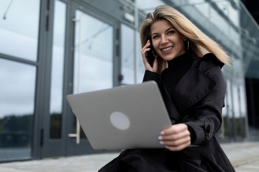 blonde middle-aged businesswoman works online on a laptop and speaks on a mobile phone against the backdrop of an office building.