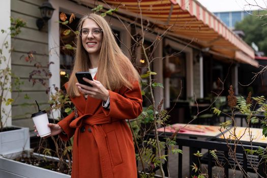 a young woman with a cup of coffee in her hands and a mobile phone looks into the distance with a smile, waiting for a young man on a date.