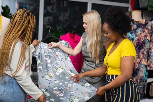 A dark-skinned expeditor shows a customer a new model of dress with a floral pattern. A blonde woman picks out a dress for summer at a tailor's shop.