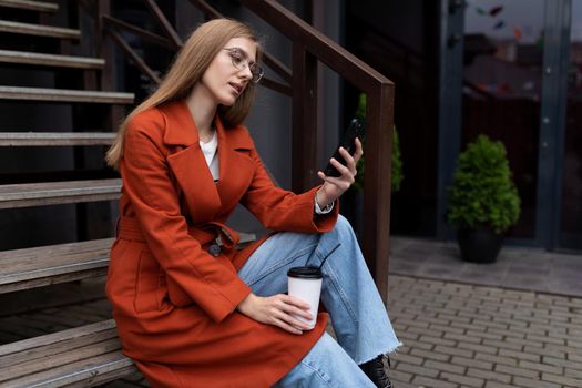 young stylish woman sits on the stairs next to a popular cafe with a cup of coffee and watches a mobile phone.