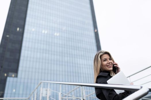 Business woman blonde against the backdrop of an office skyscraper expired with a mobile phone and a laptop.