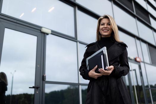 Middle-aged business woman with a laptop and a notepad at the entrance to an office building looking at the camera with a smile.