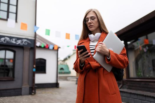 young woman in an autumn orange coat with a laptop and a mobile phone on the background of the city.