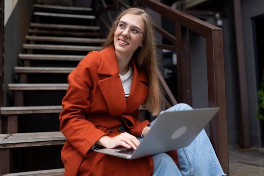 young woman working on a laptop online sitting on a flight of stairs next to a cafe smiling looking away.
