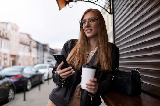 young woman with a cup of coffee by a mobile phone with a smile looks into the distance against the backdrop of the city.