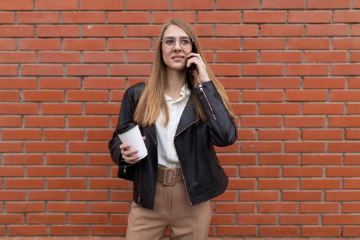 young female student talking on a mobile phone with a cup of coffee in her hands against the background of an orange brick wall.