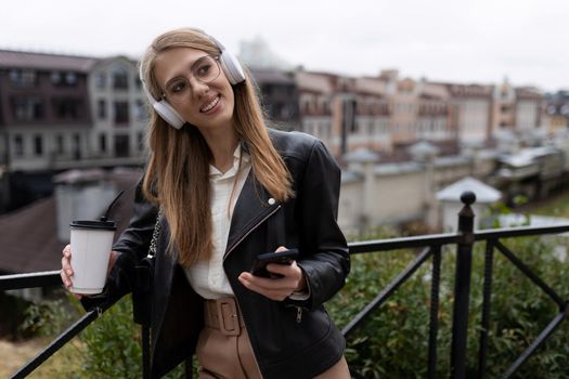 portrait of a cheerful and happy young woman with a mobile phone and a cup of coffee in her hands listening to music through over-ear headphones with a smile on her face.