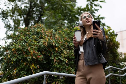 young female student listening to music in headphones with a cup of coffee and a phone in her hand in the city.