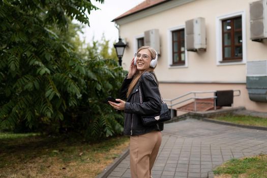 cheerful naughty young blonde woman walks around the city with a mobile phone and listens to music in headphones while dancing.