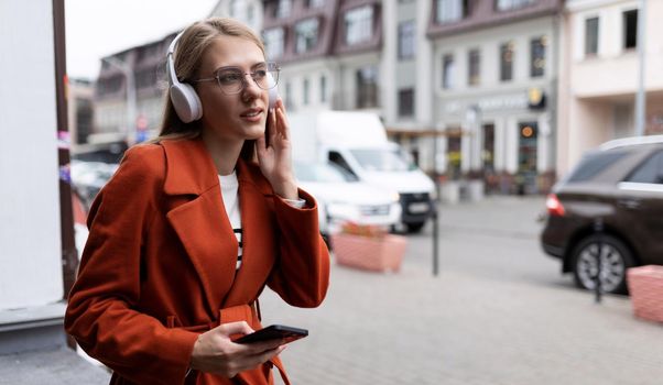 a young woman listens to music in headphones with a phone in her hands against the backdrop of city cafes.