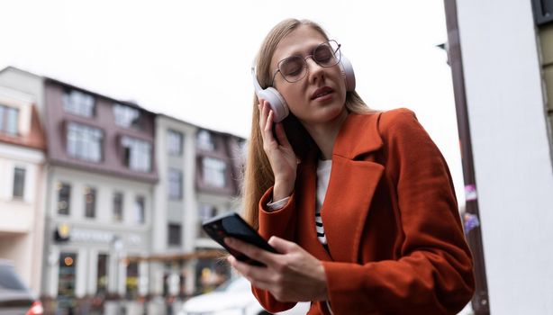 young blonde woman with a mobile phone in her hands listens to music on headphones against the backdrop of the urban landscape.