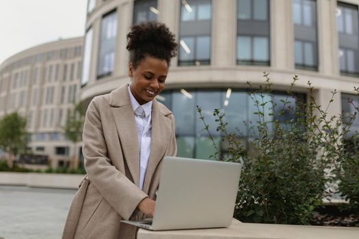 young black woman working on a laptop online against the backdrop of the city with a smile on her face.