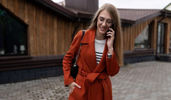 young beautiful woman in a coat talking on a mobile phone against the backdrop of a city building with a smile on her face.