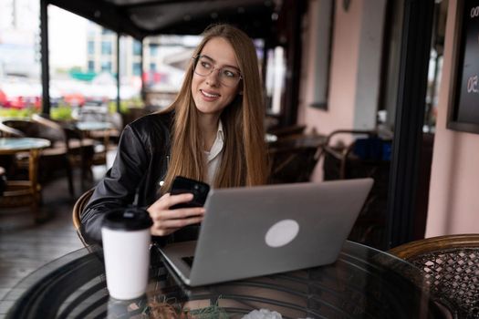 young woman freelancer drinking coffee and working online on a laptop in a city cafe with a smile looking at the camera.