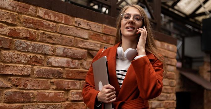 young woman rushing to work with a smile on her face with a laptop under her arm talking on a mobile phone against the backdrop of the city wall of the building.